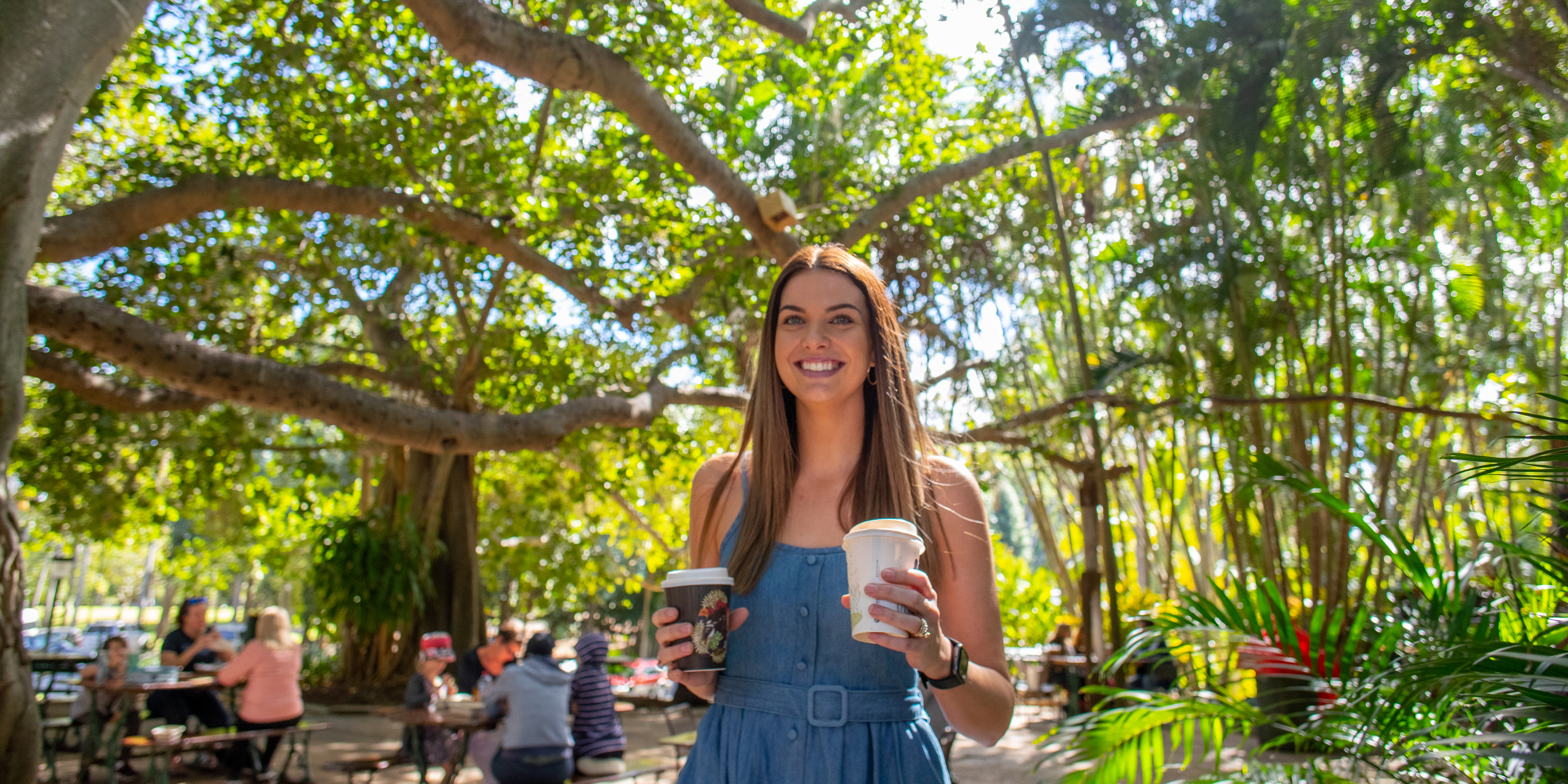 Young woman holding coffee with people sitting at chairs and tables under a giant fig tree