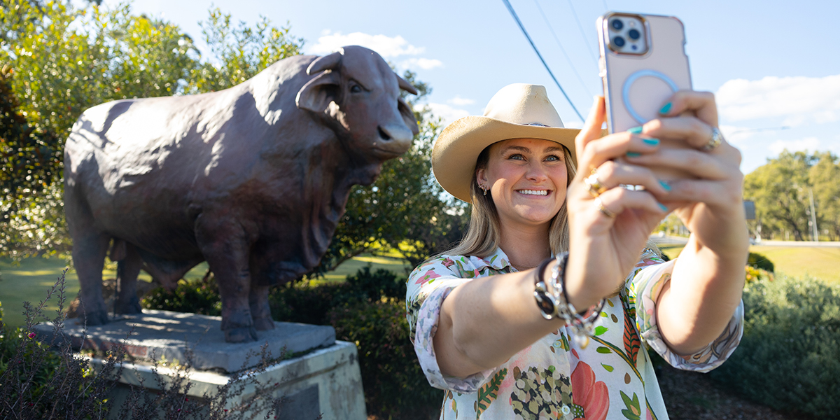 Young, beautiful woman wearing a cowboy hat, smiling, while taking a selfie with a life-size bull statue