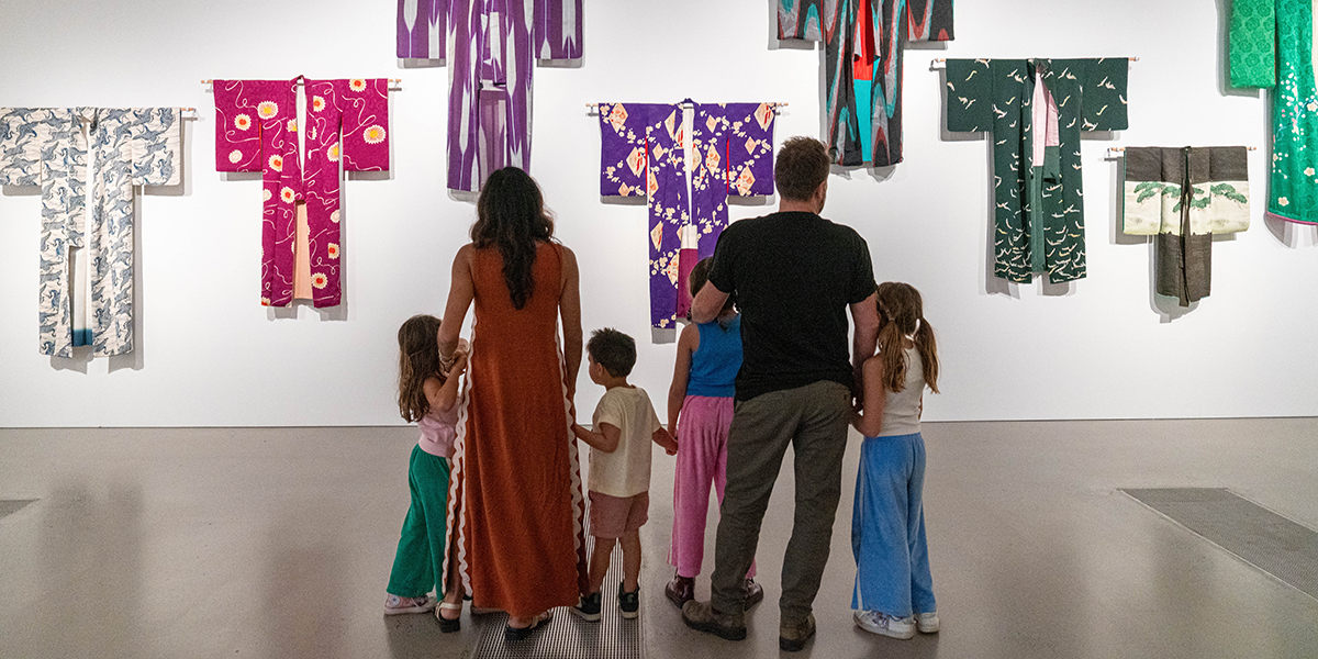 Family of six with their back to the camera looking at artwork of kimonos hung neatly against a white wall.