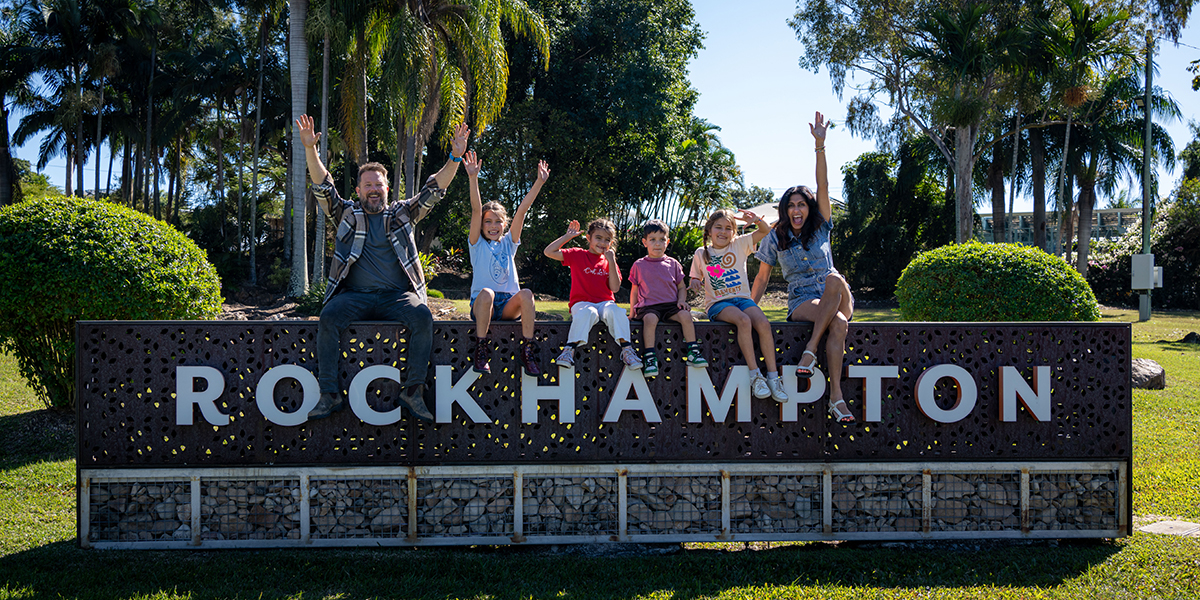 Family of six sitting on a giant sign that reads Rockhampton smiling and raising their arms in the air in excitement.