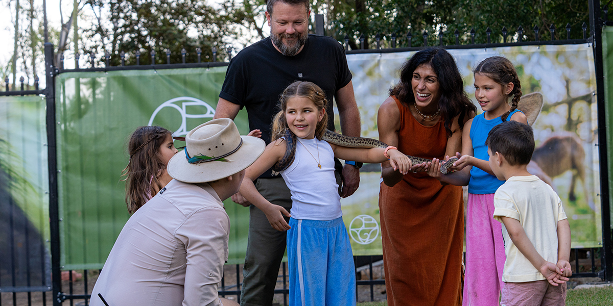 Young girl nursing a python around her neck while her family surrounds her learning about reptiles from the zoo keeper.