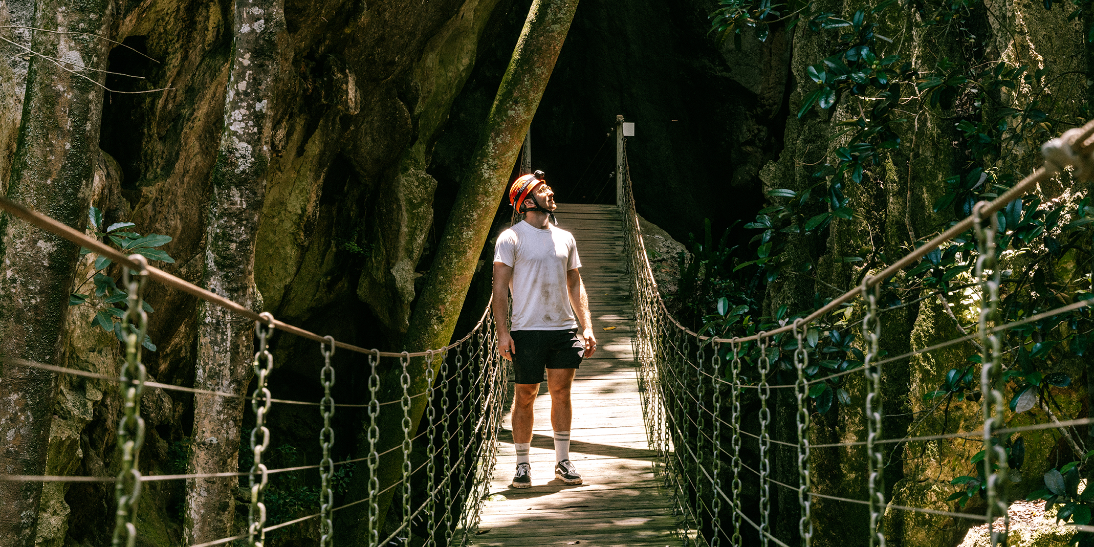 Young man wearing helmet stands on a walking bridge with cave entrance in the background