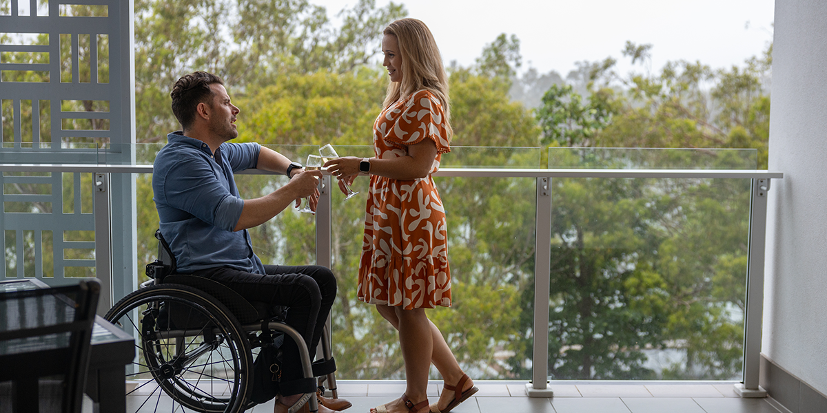 A man in a wheelchair and an able-bodied woman standing beside him clinking wine glasses as they overlook the river from their hotel balcony