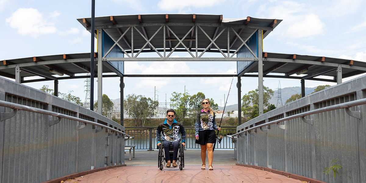 A man in a wheelchair and an able-bodied woman using the ramp accessible land based fishing platform carrying a fishing rod