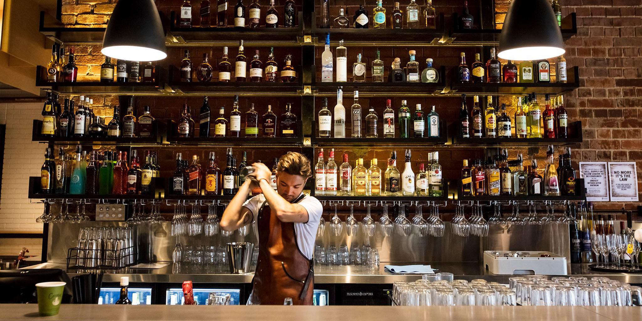 Bartender shaking up a cocktail with a wall of spirits shelved behind him at Headricks Lane.