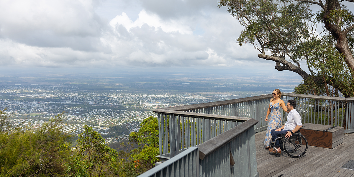 A man in a wheelchair and a woman on the boardwalk overlooking the Rockhampton City from Mount Archer National Park via the accessible nature trail