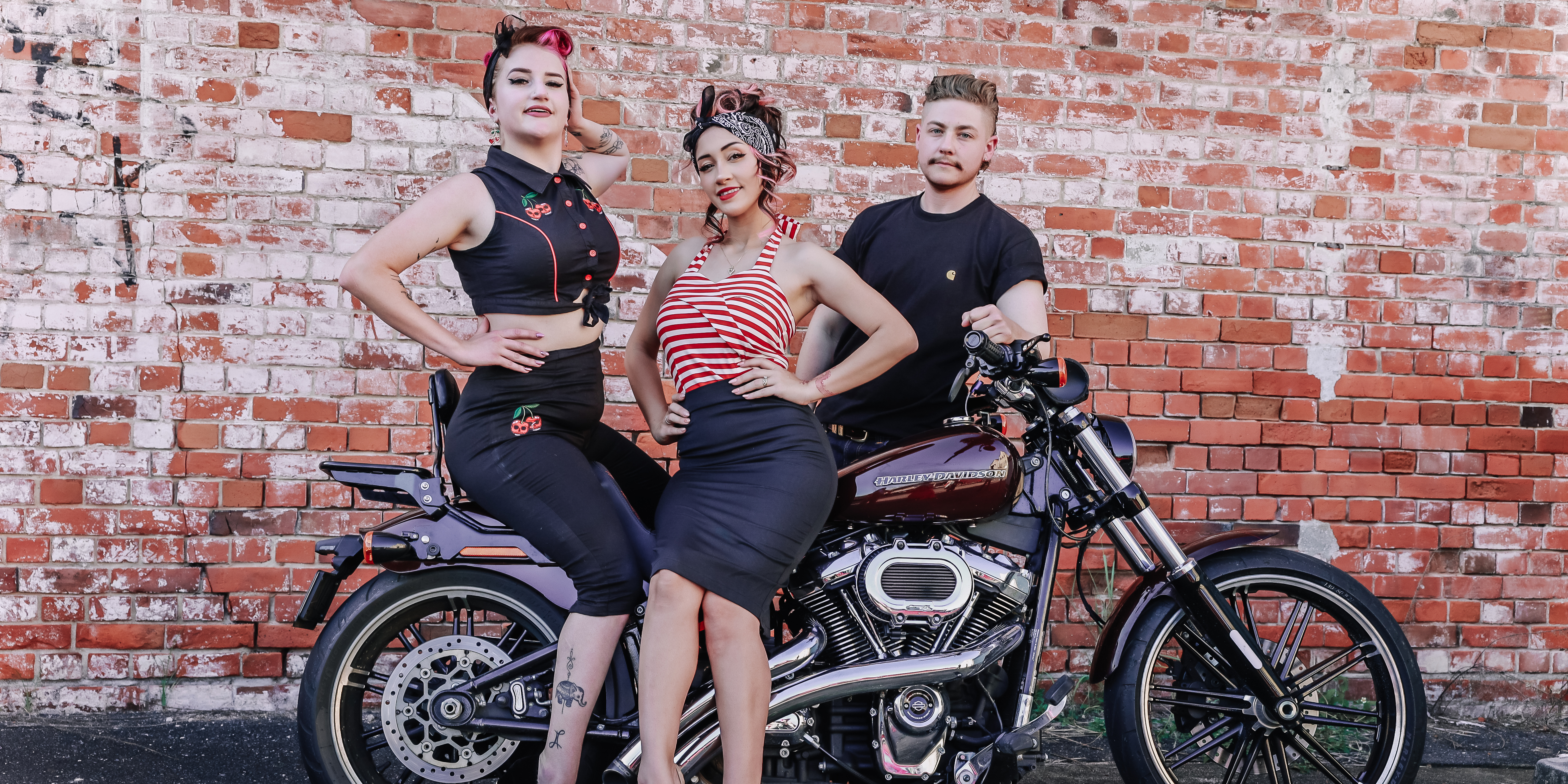 Two women dressed in pinup clothing sitting on a motorcycle and a gentleman standing behind