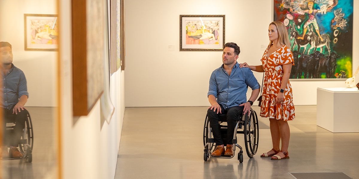 A man in a wheelchair with a woman standing beside him admiring the artwork on the wall in the Rockhampton Museum of Art gallery