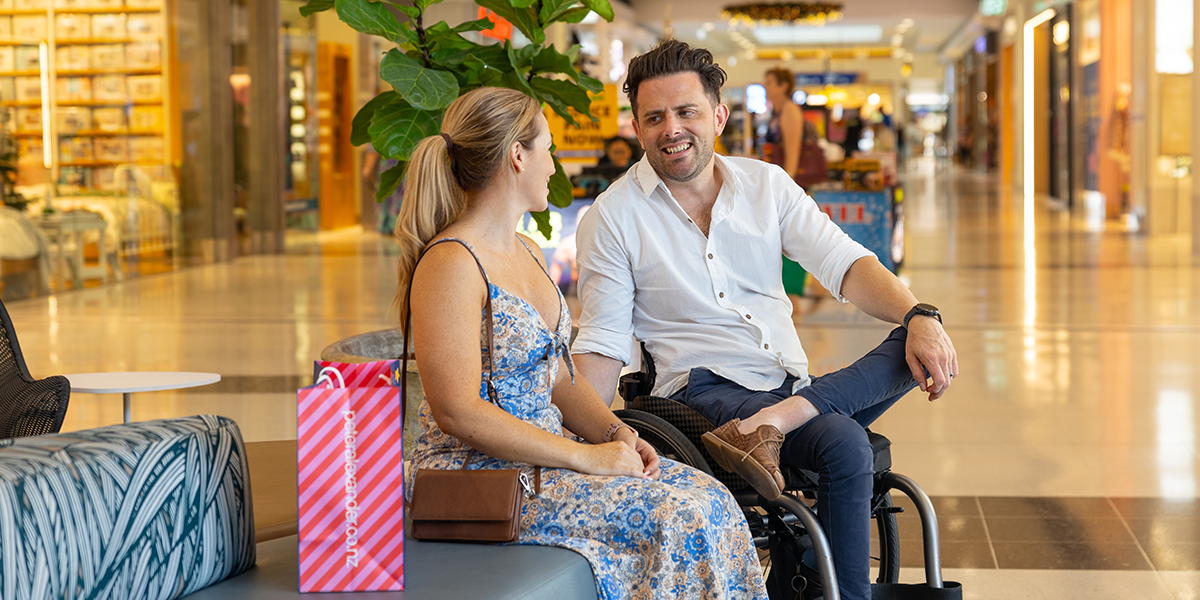A woman sitting with a shopping bag on a shopping centre lounge and a man in a wheelchair beside her. There are people in the background as well as specialty stores from well-known brands