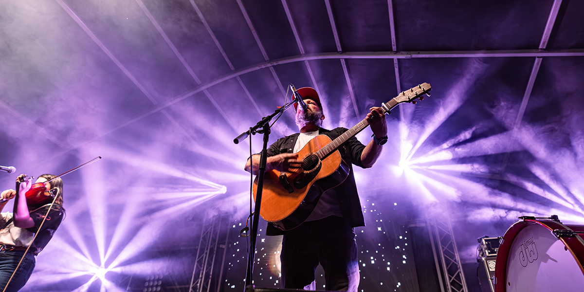 An upwards shot of Tom Busby playing guitar and singing on stage at the Rockhampton River Festival with purple lights illuminating the background.