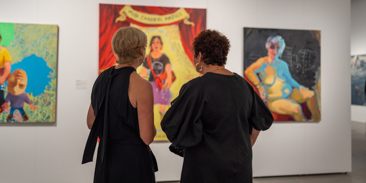Two women with their back facing the camera looking at painted artwork on a gallery wall.