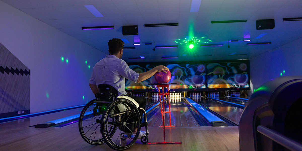 A man in a wheelchair holding a bowling ball on a bowl ramp in a bowling alley lit with disco lights