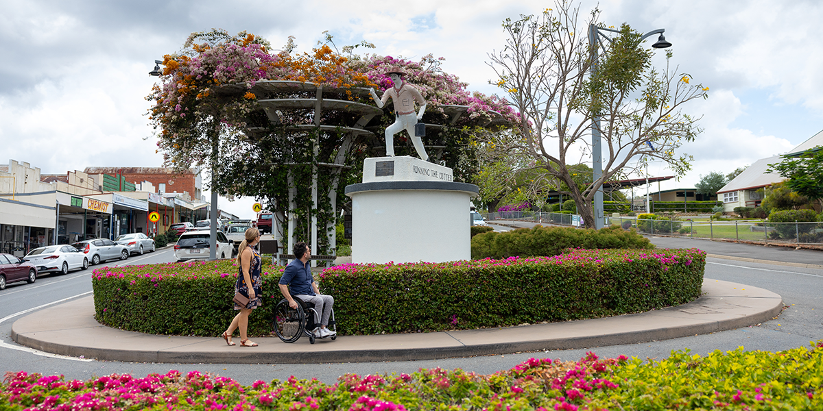 A man  in a wheelchair and an able-bodied woman exploring a statue of a man running a mining cutter with a background of pink flowers