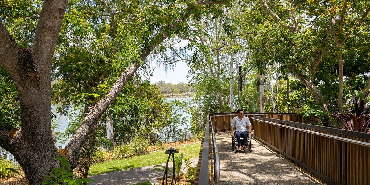 A man in a wheelchair using an access ramp in the shade of the leafy trees along the river